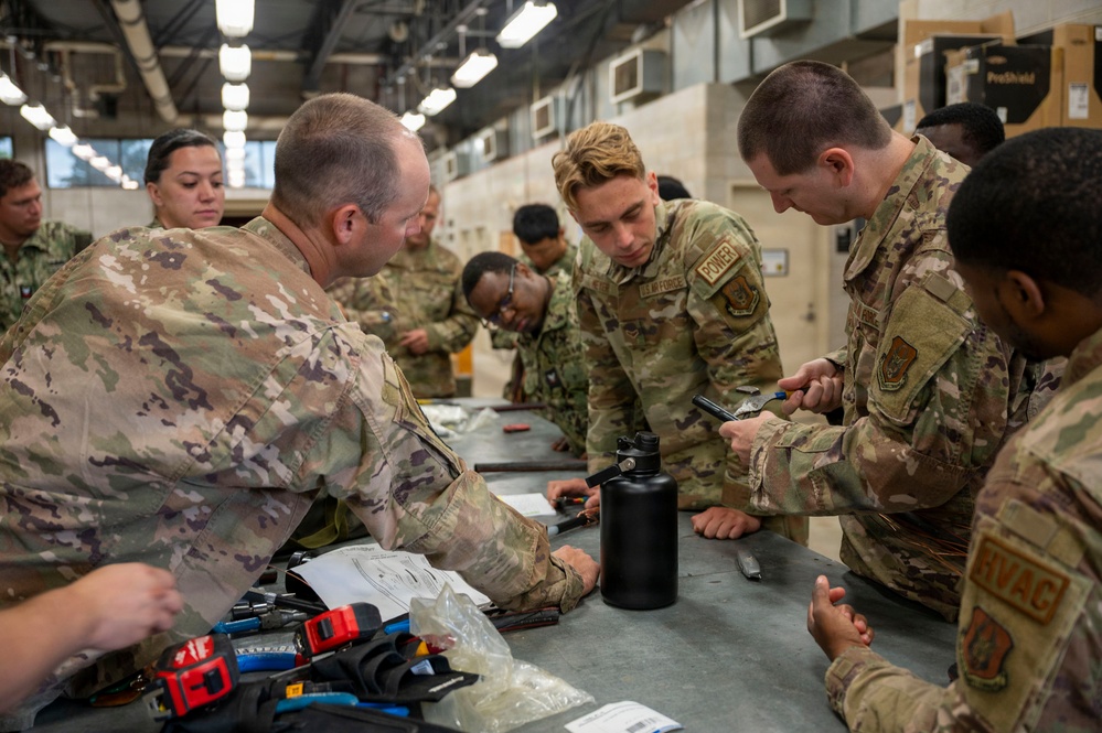 A group of military personnel in OCP (Operational Camouflage Pattern) uniforms are gathered around a workbench in what appears to be a hangar or maintenance facility. They appear to be engaged in a hands-on training exercise involving tools and technical components. At least one patch reading 'U.S. AIR FORCE' and another reading 'HVAC' are visible, suggesting Air Force technical training.