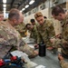 A group of military personnel in OCP (Operational Camouflage Pattern) uniforms are gathered around a workbench in what appears to be a hangar or maintenance facility. They appear to be engaged in a hands-on training exercise involving tools and technical components. At least one patch reading 'U.S. AIR FORCE' and another reading 'HVAC' are visible, suggesting Air Force technical training.