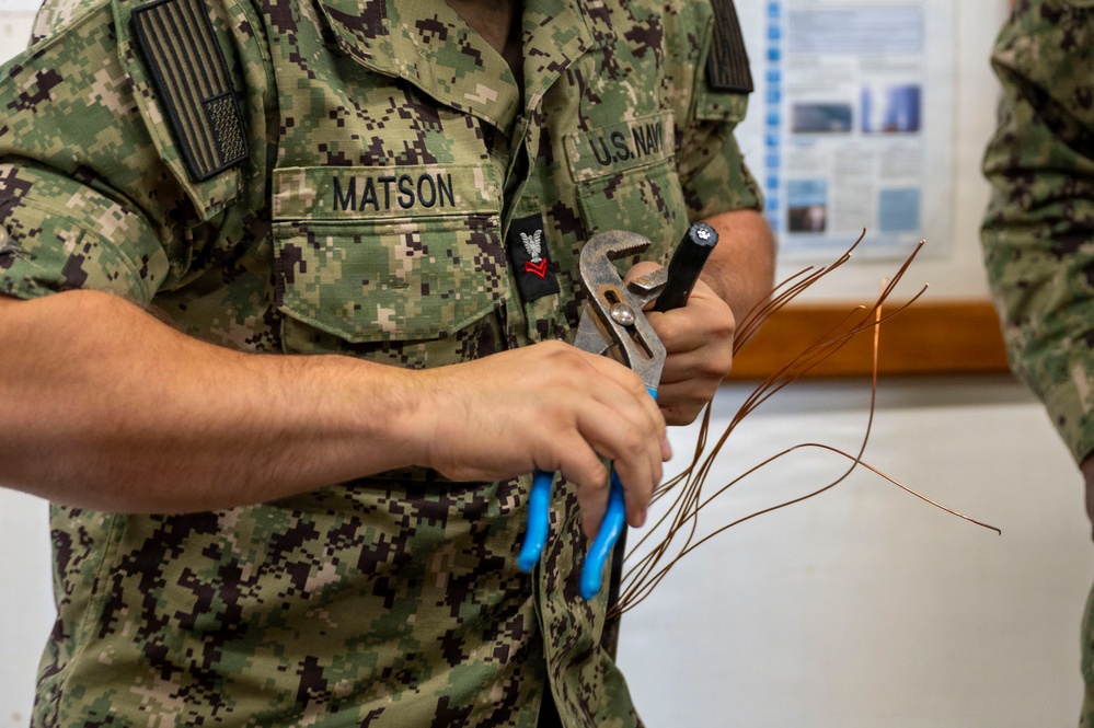 A U.S. Navy sailor named Matson, wearing NWU (Navy Working Uniform) digital camouflage, is holding tools and copper wiring. The sailor appears to be engaged in an electrical or technical training exercise, holding both pliers with blue handles and an adjustable wrench along with stripped copper wire strands. A second uniformed individual is partially visible on the right side of the frame.