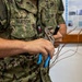 A U.S. Navy sailor named Matson, wearing NWU (Navy Working Uniform) digital camouflage, is holding tools and copper wiring. The sailor appears to be engaged in an electrical or technical training exercise, holding both pliers with blue handles and an adjustable wrench along with stripped copper wire strands. A second uniformed individual is partially visible on the right side of the frame.
