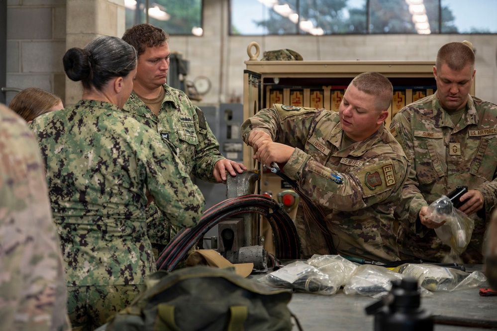 Military personnel from multiple branches, including the U.S. Navy and U.S. Air Force, are gathered around a workbench in what appears to be a maintenance or training facility. A U.S. Air Force member wearing an 'ELEC' (Electrical) patch and name tape 'HARPER' is demonstrating or performing work on a piece of equipment. The group appears to be engaged in a joint-service training or maintenance exercise.