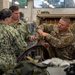 Military personnel from multiple branches, including the U.S. Navy and U.S. Air Force, are gathered around a workbench in what appears to be a maintenance or training facility. A U.S. Air Force member wearing an 'ELEC' (Electrical) patch and name tape 'HARPER' is demonstrating or performing work on a piece of equipment. The group appears to be engaged in a joint-service training or maintenance exercise.
