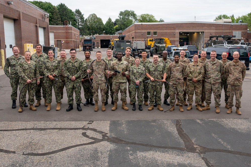 A large group of approximately 22 military personnel in camouflage uniforms pose for a formal group photograph in front of a brick facility with garage bays. The group includes members wearing both Navy Working Uniform (NWU) digital blue-gray camouflage and Army/Air Force OCP (Operational Camouflage Pattern) uniforms, suggesting a joint-service gathering. Several personnel are holding what appear to be cables or wiring, possibly indicating a training or technical exercise context.