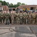 A large group of approximately 22 military personnel in camouflage uniforms pose for a formal group photograph in front of a brick facility with garage bays. The group includes members wearing both Navy Working Uniform (NWU) digital blue-gray camouflage and Army/Air Force OCP (Operational Camouflage Pattern) uniforms, suggesting a joint-service gathering. Several personnel are holding what appear to be cables or wiring, possibly indicating a training or technical exercise context.