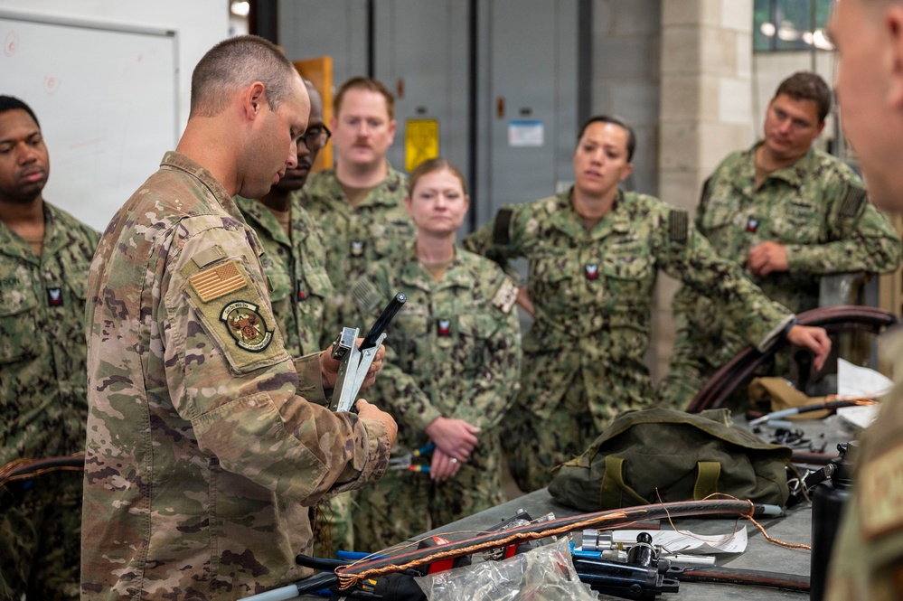 A military training or briefing session is taking place indoors, where an Air Force instructor in OCP (Operational Camouflage Pattern) uniform is demonstrating a tool or piece of equipment to a group of Navy personnel in NWU (Navy Working Uniform) camouflage. The group is gathered around a table covered with various tools, cables, and equipment. The setting appears to be a maintenance bay or training facility.