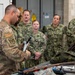 A military training or briefing session is taking place indoors, where an Air Force instructor in OCP (Operational Camouflage Pattern) uniform is demonstrating a tool or piece of equipment to a group of Navy personnel in NWU (Navy Working Uniform) camouflage. The group is gathered around a table covered with various tools, cables, and equipment. The setting appears to be a maintenance bay or training facility.