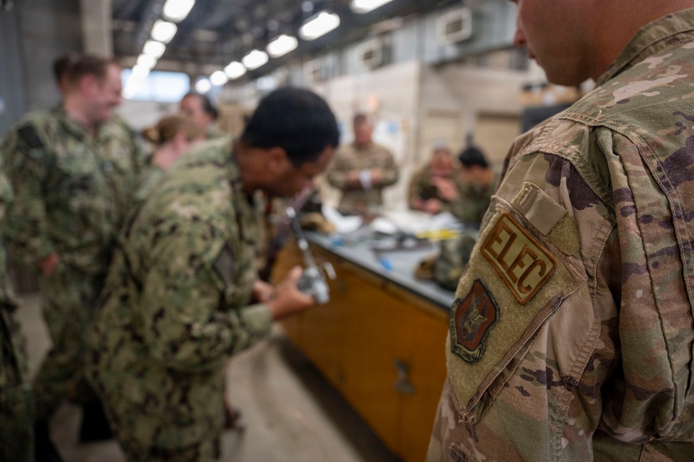 Military personnel from what appears to be multiple branches are gathered around a workbench in an indoor facility, engaged in hands-on training or equipment maintenance. The foreground shows a service member in OCP (Operational Camouflage Pattern) uniform with an 'ELEC' (Electrician) patch and a unit patch visible on the sleeve. The background shows additional personnel in Navy Working Uniform (NWU) camouflage pattern.