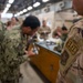 Military personnel from what appears to be multiple branches are gathered around a workbench in an indoor facility, engaged in hands-on training or equipment maintenance. The foreground shows a service member in OCP (Operational Camouflage Pattern) uniform with an 'ELEC' (Electrician) patch and a unit patch visible on the sleeve. The background shows additional personnel in Navy Working Uniform (NWU) camouflage pattern.