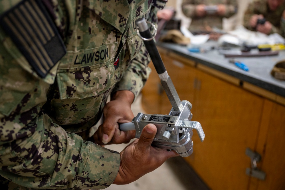 A U.S. Navy service member with the name tape 'LAWSON' is handling a piece of military equipment, likely a radiation detection or measurement device, in what appears to be an indoor training or laboratory environment. The individual is wearing Navy Working Uniform (NWU) Type III digital camouflage. Additional personnel and equipment are visible in the background on a workbench.