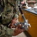 A U.S. Navy service member with the name tape 'LAWSON' is handling a piece of military equipment, likely a radiation detection or measurement device, in what appears to be an indoor training or laboratory environment. The individual is wearing Navy Working Uniform (NWU) Type III digital camouflage. Additional personnel and equipment are visible in the background on a workbench.