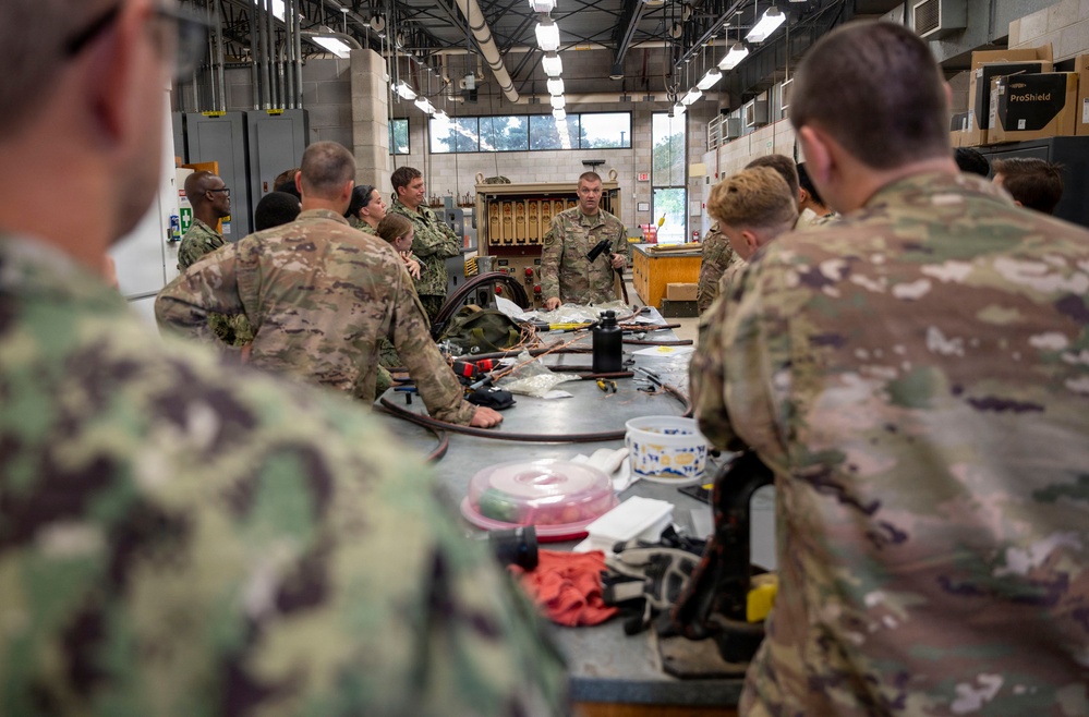 Military personnel in camouflage uniforms are gathered around a large workbench in what appears to be a maintenance or training facility. A central figure, likely an instructor, is holding a component and conducting a briefing or demonstration. The setting suggests a technical training session involving equipment maintenance or familiarization.