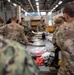 Military personnel in camouflage uniforms are gathered around a large workbench in what appears to be a maintenance or training facility. A central figure, likely an instructor, is holding a component and conducting a briefing or demonstration. The setting suggests a technical training session involving equipment maintenance or familiarization.
