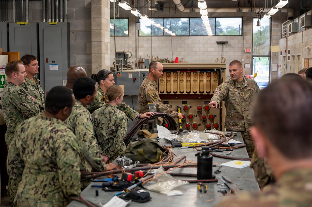 Military personnel from multiple branches, including U.S. Navy (NWU Type III camouflage) and U.S. Army (OCP/MultiCam), are gathered in an industrial workshop or training facility. Two Army soldiers appear to be instructing the group near a large tan-colored equipment rack with multiple switches and controls. A workbench in the foreground is covered with cables, wiring, tools, and various components suggesting hands-on technical training.