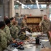 Military personnel from multiple branches, including U.S. Navy (NWU Type III camouflage) and U.S. Army (OCP/MultiCam), are gathered in an industrial workshop or training facility. Two Army soldiers appear to be instructing the group near a large tan-colored equipment rack with multiple switches and controls. A workbench in the foreground is covered with cables, wiring, tools, and various components suggesting hands-on technical training.