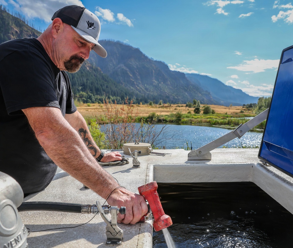 A bearded man wearing a black t-shirt and gray trucker cap is operating a red water nozzle/hose fitting, directing water into a tank or trough on what appears to be a utility vehicle or water tender. The setting is a scenic mountain valley with a river or pond, open meadows, and partially burned hillsides visible in the background. The image appears to document a water-filling or water-hauling operation in a rural or wildfire-affected area.