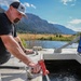 A bearded man wearing a black t-shirt and gray trucker cap is operating a red water nozzle/hose fitting, directing water into a tank or trough on what appears to be a utility vehicle or water tender. The setting is a scenic mountain valley with a river or pond, open meadows, and partially burned hillsides visible in the background. The image appears to document a water-filling or water-hauling operation in a rural or wildfire-affected area.