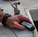 A person is holding a Fish Hawk TD water temperature and depth probe device, which displays a reading of 64.3 degrees on its LCD screen. The individual is wearing a black sport watch and appears to be on a boat, resting the device on the gunwale or deck edge. The device is a fishing tool used to measure water temperature at various depths.