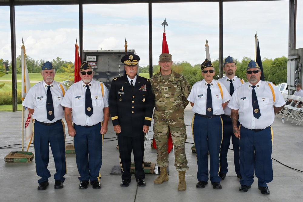 A group of seven men pose for a formal photograph at what appears to be a military or veterans' ceremony. Two individuals are in active military uniforms, while five others wear what appear to be American Legion or similar veterans' organization dress uniforms consisting of white shirts, blue trousers, and blue garrison caps. The setting is an open-air pavilion or hangar with color guard flags visible in the background.