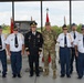A group of seven men pose for a formal photograph at what appears to be a military or veterans' ceremony. Two individuals are in active military uniforms, while five others wear what appear to be American Legion or similar veterans' organization dress uniforms consisting of white shirts, blue trousers, and blue garrison caps. The setting is an open-air pavilion or hangar with color guard flags visible in the background.