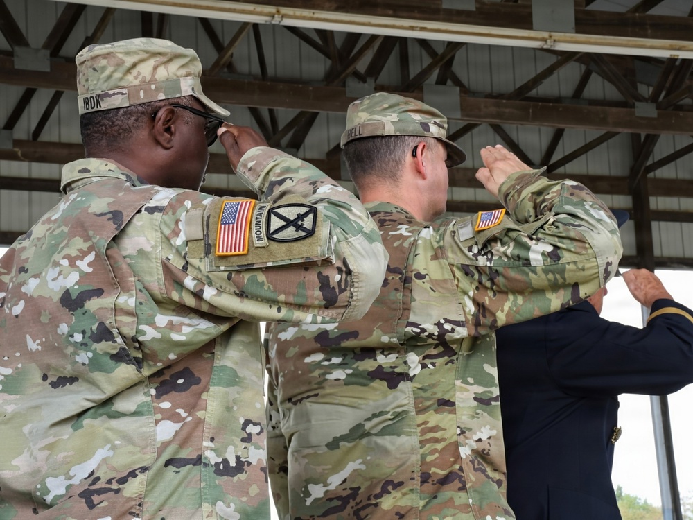 Three military personnel in U.S. Army OCP (Operational Camouflage Pattern) uniforms are rendering a salute, likely during a formal military ceremony. Two soldiers are facing away from the camera, with name tapes and unit patches visible on their uniforms. A third individual in dress blues is partially visible on the right edge of the frame.