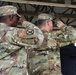 Three military personnel in U.S. Army OCP (Operational Camouflage Pattern) uniforms are rendering a salute, likely during a formal military ceremony. Two soldiers are facing away from the camera, with name tapes and unit patches visible on their uniforms. A third individual in dress blues is partially visible on the right edge of the frame.