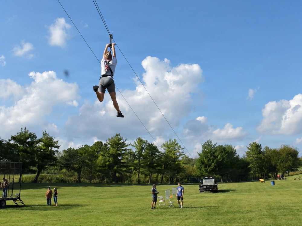 A person is riding a zipline across an open grassy field, suspended high above the ground while gripping a handle attached to the cable. Several spectators stand on the lawn below watching the activity. The setting appears to be an outdoor recreational or camp facility on a sunny day with scattered clouds.