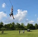 A person is riding a zipline across an open grassy field, suspended high above the ground while gripping a handle attached to the cable. Several spectators stand on the lawn below watching the activity. The setting appears to be an outdoor recreational or camp facility on a sunny day with scattered clouds.
