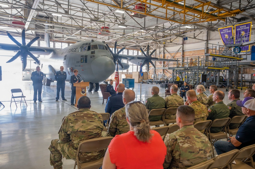 A military ceremony is taking place inside an aircraft hangar, with a speaker at a podium addressing an audience of seated military personnel and civilians. A large US Air Force C-130 Hercules transport aircraft serves as the backdrop, marked with '934 AW' indicating the 934th Airlift Wing. A banner on the right side of the hangar also references the 934th Airlift Wing.