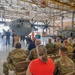A military ceremony is taking place inside an aircraft hangar, with a speaker at a podium addressing an audience of seated military personnel and civilians. A large US Air Force C-130 Hercules transport aircraft serves as the backdrop, marked with '934 AW' indicating the 934th Airlift Wing. A banner on the right side of the hangar also references the 934th Airlift Wing.