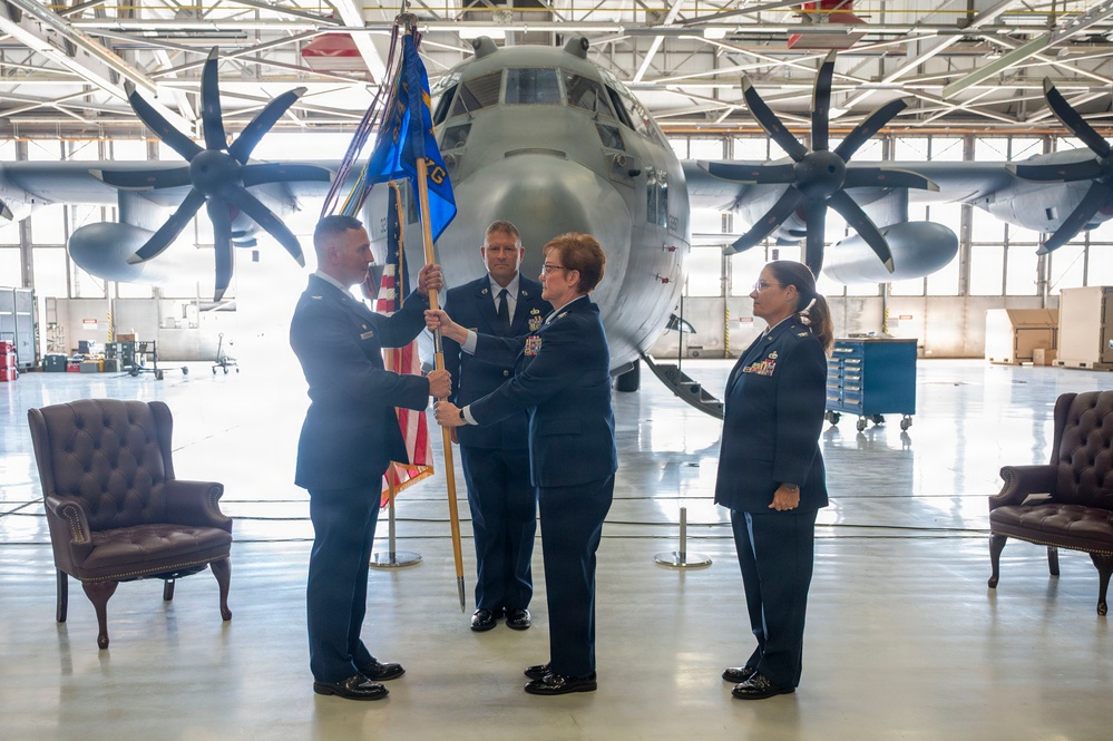 A military change of command ceremony is taking place inside an aircraft hangar, with a large turboprop military transport aircraft (appearing to be a C-130) visible in the background. Four uniformed U.S. Air Force personnel are participating, with two individuals jointly holding a blue unit guidon/flag, which is the central symbolic act of a change of command. Two leather chairs are positioned on either side of the ceremony space, consistent with formal military protocol.