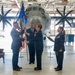 A military change of command ceremony is taking place inside an aircraft hangar, with a large turboprop military transport aircraft (appearing to be a C-130) visible in the background. Four uniformed U.S. Air Force personnel are participating, with two individuals jointly holding a blue unit guidon/flag, which is the central symbolic act of a change of command. Two leather chairs are positioned on either side of the ceremony space, consistent with formal military protocol.