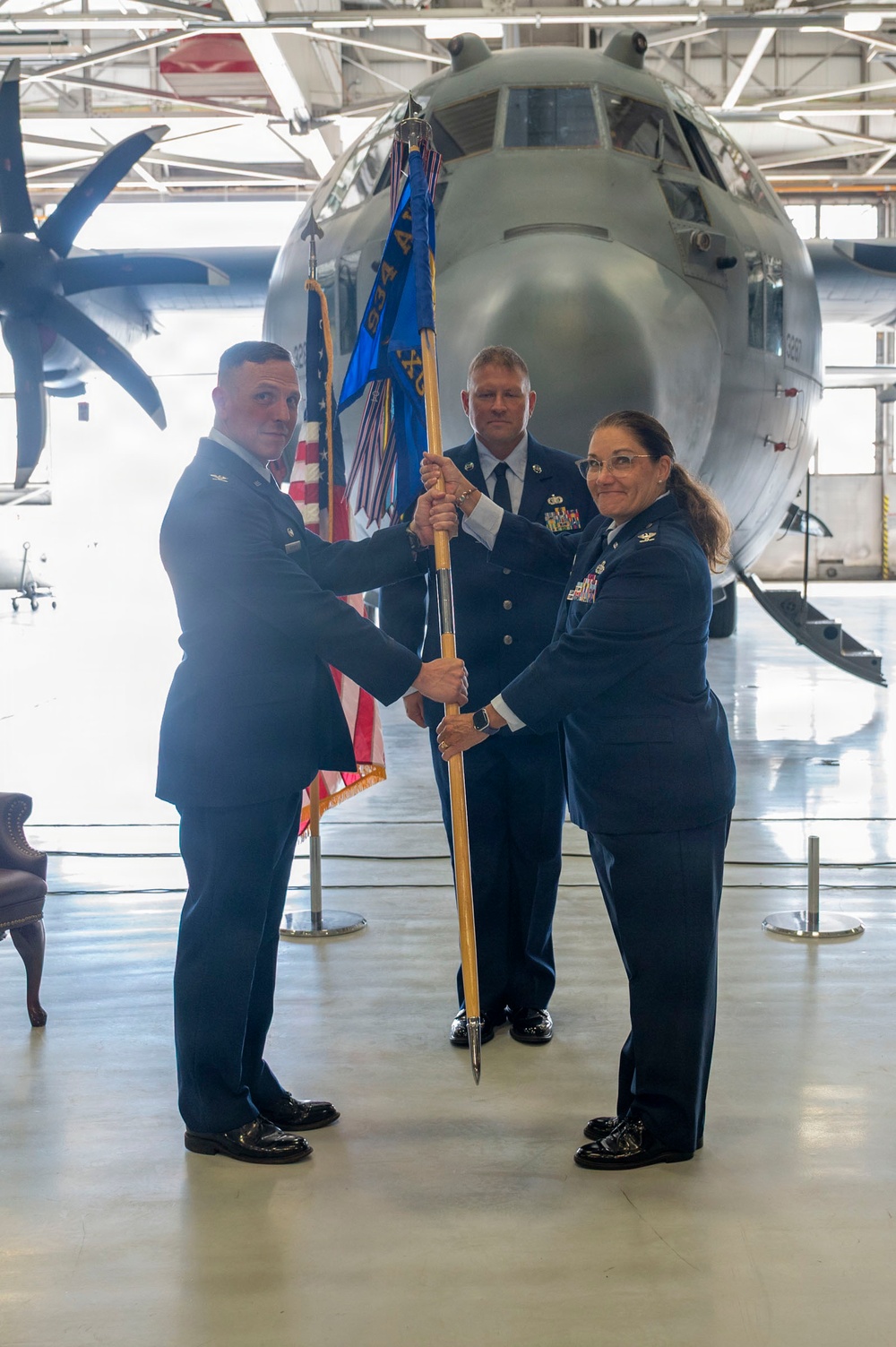 A military change of command ceremony taking place inside an aircraft hangar. Two officers in US Air Force dress blue uniforms are exchanging a unit guidon (flag on a staff), with a third officer standing at attention in the background. A large military transport aircraft, appearing to be a C-130, is visible behind the participants.