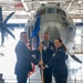 A military change of command ceremony taking place inside an aircraft hangar. Two officers in US Air Force dress blue uniforms are exchanging a unit guidon (flag on a staff), with a third officer standing at attention in the background. A large military transport aircraft, appearing to be a C-130, is visible behind the participants.