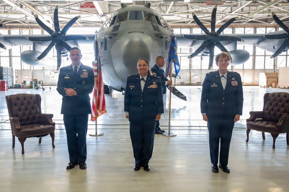 Three U.S. Air Force personnel in dress blue uniforms are posed in a military hangar in front of a large turboprop aircraft, likely a C-130J Super Hercules. The scene appears to be a formal military ceremony, possibly a change of command or promotion, indicated by the presence of a unit guidon, an American flag, and ceremonial chairs. A fourth service member is visible in the background holding the guidon staff.