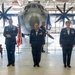 Three U.S. Air Force personnel in dress blue uniforms are posed in a military hangar in front of a large turboprop aircraft, likely a C-130J Super Hercules. The scene appears to be a formal military ceremony, possibly a change of command or promotion, indicated by the presence of a unit guidon, an American flag, and ceremonial chairs. A fourth service member is visible in the background holding the guidon staff.