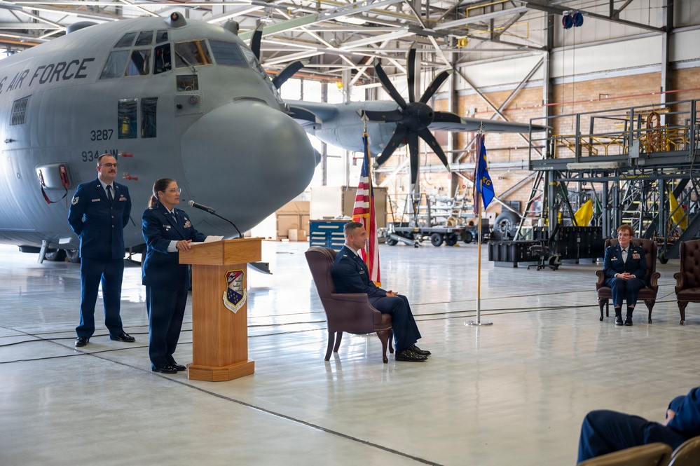 A military ceremony is taking place inside an aircraft hangar, with a female Air Force officer speaking at a podium bearing a unit insignia. A large C-130 Hercules transport aircraft (tail number 3287, marked '934 AW' and 'US AIR FORCE') serves as the backdrop. The setting and seating arrangement suggest this is a formal change of command or award ceremony.