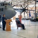 A military ceremony is taking place inside an aircraft hangar, with a female Air Force officer speaking at a podium bearing a unit insignia. A large C-130 Hercules transport aircraft (tail number 3287, marked '934 AW' and 'US AIR FORCE') serves as the backdrop. The setting and seating arrangement suggest this is a formal change of command or award ceremony.