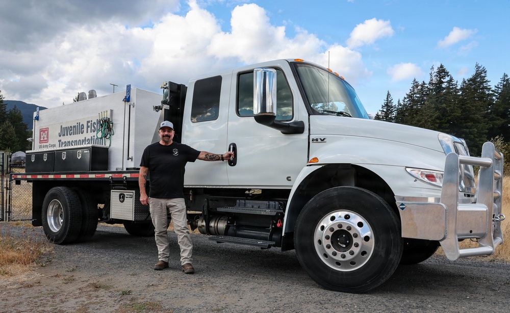 A man stands beside a large white US Army Corps of Engineers 'Juvenile Fish Transportation' truck, posing with his hand on the door handle. The truck appears to be a heavy-duty International MV series vehicle equipped with a specialized tank unit mounted on the flatbed. The setting appears to be a gravel lot near a fenced facility with forested mountains in the background.