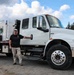 A man stands beside a large white US Army Corps of Engineers 'Juvenile Fish Transportation' truck, posing with his hand on the door handle. The truck appears to be a heavy-duty International MV series vehicle equipped with a specialized tank unit mounted on the flatbed. The setting appears to be a gravel lot near a fenced facility with forested mountains in the background.