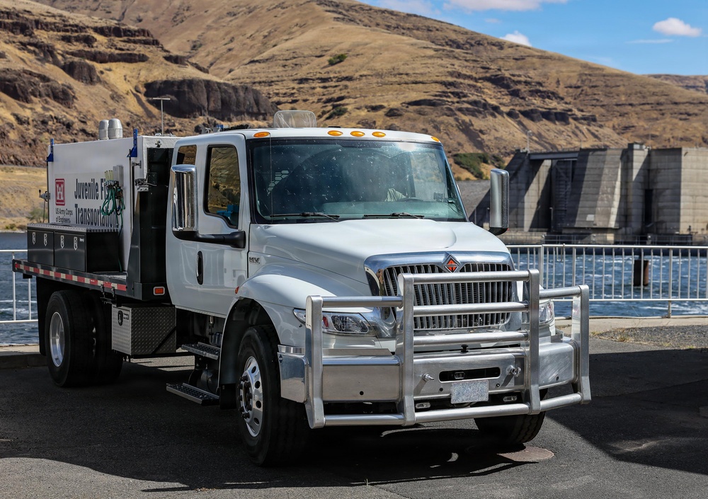 A white US Army Corps of Engineers 'Juvenile Fish Transportation' truck is parked near a dam or water control structure. The International-brand medium-duty truck features a chrome grille guard and is equipped with a specialized tank/transport unit on the flatbed. The setting appears to be a river dam facility in an arid, hilly landscape.
