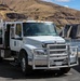 A white US Army Corps of Engineers 'Juvenile Fish Transportation' truck is parked near a dam or water control structure. The International-brand medium-duty truck features a chrome grille guard and is equipped with a specialized tank/transport unit on the flatbed. The setting appears to be a river dam facility in an arid, hilly landscape.