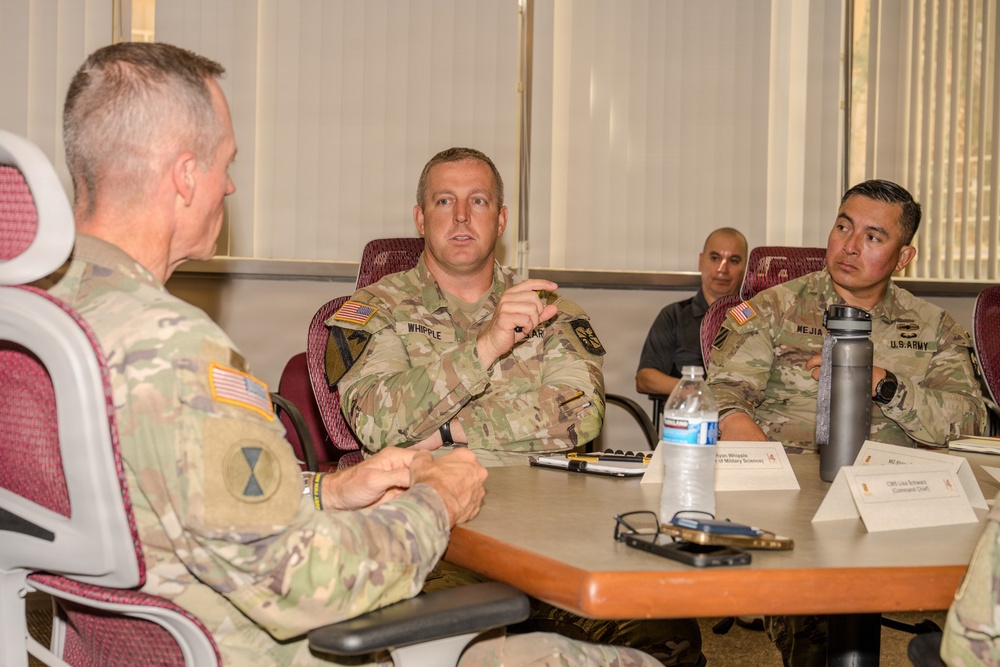 A group of U.S. Army personnel are seated around a conference table engaged in a discussion or briefing. The central figure, identified by a name tag as 'WHIPPLE,' appears to be speaking or presenting, while others listen attentively. Name placards are visible on the table, indicating a formal meeting setting.