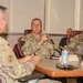 A group of U.S. Army personnel are seated around a conference table engaged in a discussion or briefing. The central figure, identified by a name tag as 'WHIPPLE,' appears to be speaking or presenting, while others listen attentively. Name placards are visible on the table, indicating a formal meeting setting.