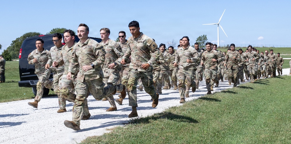 A large formation of military personnel in camouflage uniforms (OCP/MultiCam pattern) are conducting a group run along a gravel path in a rural outdoor setting. The soldiers appear to be in a physical training or unit run formation, moving in an organized group. The setting includes green grass, trees, a black SUV, and a wind turbine visible in the background under clear blue skies.