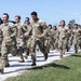A large formation of military personnel in camouflage uniforms (OCP/MultiCam pattern) are conducting a group run along a gravel path in a rural outdoor setting. The soldiers appear to be in a physical training or unit run formation, moving in an organized group. The setting includes green grass, trees, a black SUV, and a wind turbine visible in the background under clear blue skies.