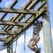 Two military personnel in camouflage uniforms are climbing ropes on an outdoor obstacle course. One soldier is near the top of the rope approaching a wooden overhead structure, while the other is lower on a separate rope. The structure includes wooden beams, vertical posts, and a cargo net, consistent with a military training facility.