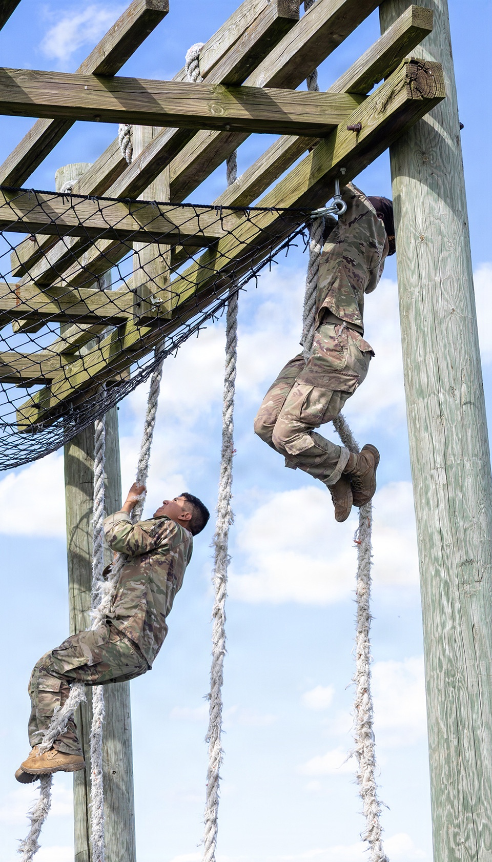 Two military personnel in camouflage uniforms are climbing ropes on an outdoor obstacle course. One soldier is near the top of the rope approaching a wooden overhead structure, while the other is lower on a separate rope. The structure includes wooden beams, vertical posts, and a cargo net, consistent with a military training facility.