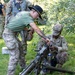 Two U.S. Army soldiers are working together on a crew-served weapon mounted on a tripod, likely during a training exercise. One soldier wearing a black cavalry Stetson hat and green t-shirt is leaning over to assist or instruct another soldier in full multicam uniform who is crouching and handling the weapon. Additional soldiers in full combat gear are visible in the background.