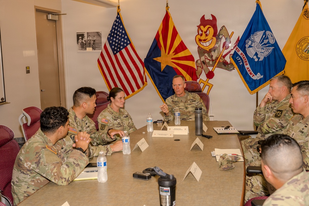 A group of U.S. Army personnel in OCP (Operational Camouflage Pattern) uniforms are seated around a conference table in what appears to be a military briefing or meeting room. A senior officer appears to be leading the discussion, with name placards visible on the table. The room displays multiple flags including the U.S. flag, Arizona state flag, United States Army flag, and an Arizona State University Sun Devils mascot cutout.