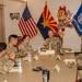 A group of U.S. Army personnel in OCP (Operational Camouflage Pattern) uniforms are seated around a conference table in what appears to be a military briefing or meeting room. A senior officer appears to be leading the discussion, with name placards visible on the table. The room displays multiple flags including the U.S. flag, Arizona state flag, United States Army flag, and an Arizona State University Sun Devils mascot cutout.